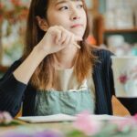 Pensive Asian florist looking away while sitting at table with hand near face and cup of hot beverage in floristry store
