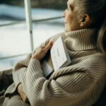 Senior woman in a cozy turtleneck embraces a book, gazing thoughtfully out the window.