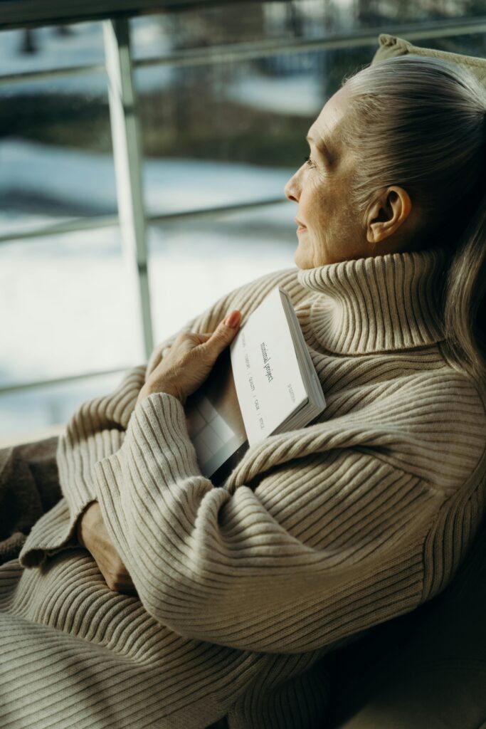 Senior woman in a cozy turtleneck embraces a book, gazing thoughtfully out the window.