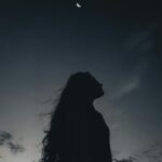 Low angle side view of silhouette of serene long haired female standing alone under night sky with moon