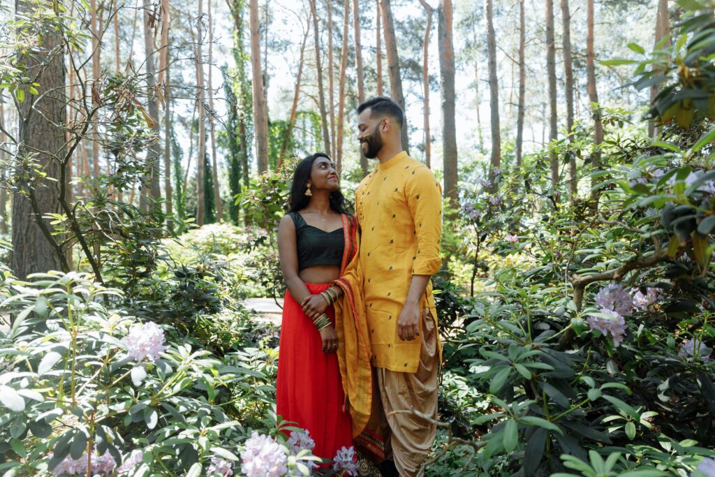 A couple in traditional Indian dress shares a loving moment in a vibrant forest setting.