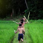 Group of children running playfully through lush green countryside, carrying sticks.