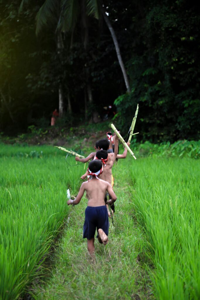 Group of children running playfully through lush green countryside, carrying sticks.