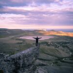 Silhouette of a man standing on a mountain edge with arms outstretched, overlooking a scenic landscape at dusk.
