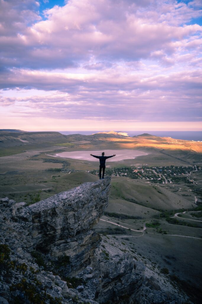 Silhouette of a man standing on a mountain edge with arms outstretched, overlooking a scenic landscape at dusk.