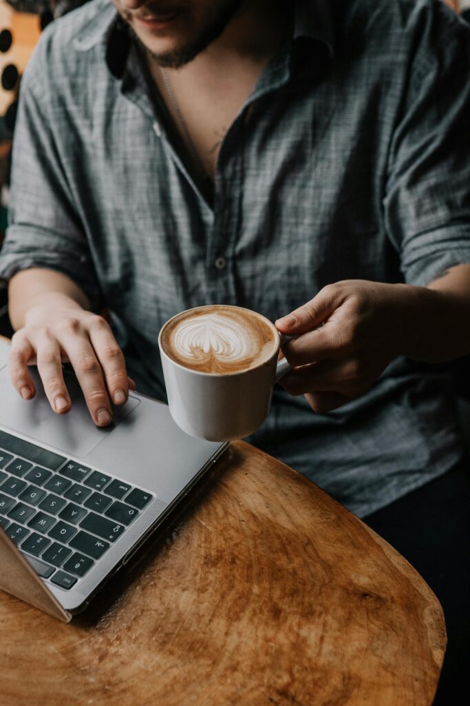 A man works on a laptop in a cozy café, holding a latte with beautiful microfoam art.