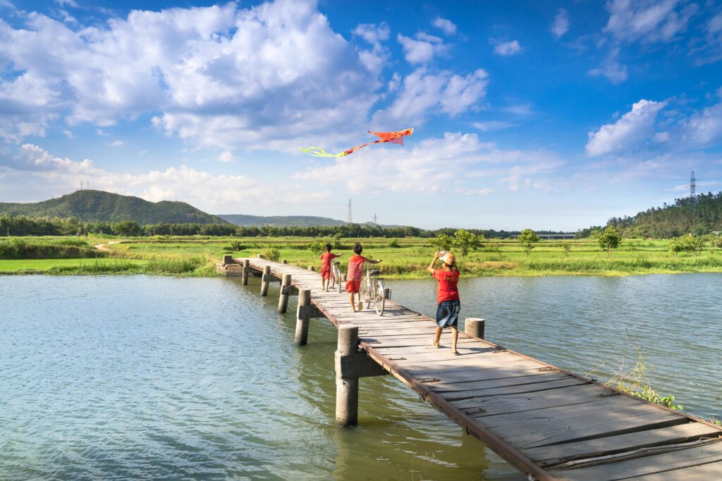 Kids enjoying a sunny day flying kites on a wooden bridge over a river in the countryside.