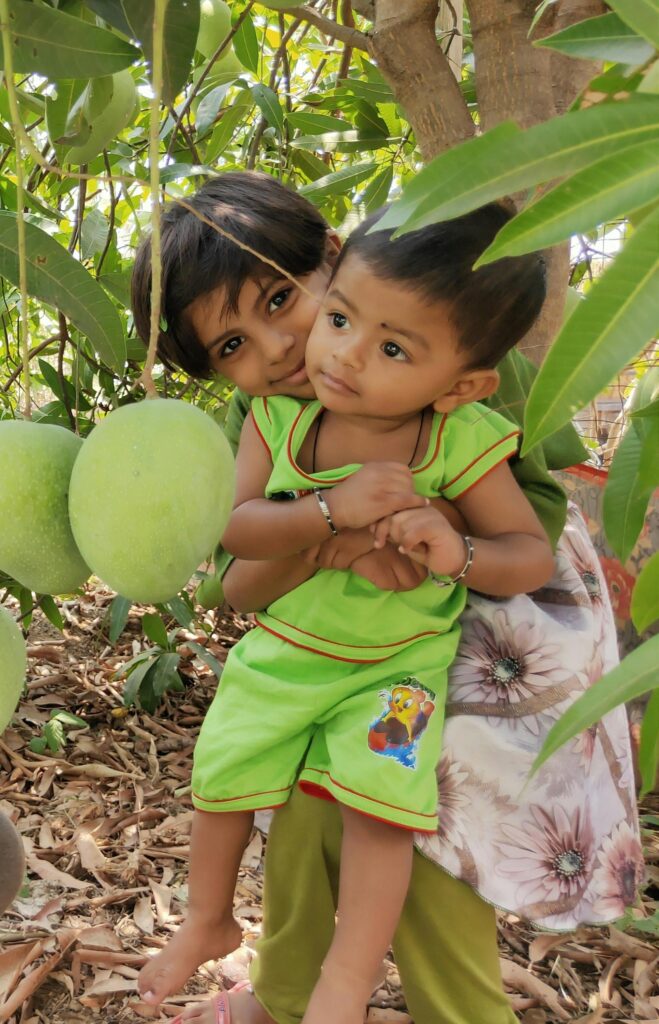 Two smiling children play under a mango tree, surrounded by lush green leaves, evoking joy and connection with nature.