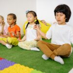 Group of diverse young children meditating in yoga poses on a green mat indoors.