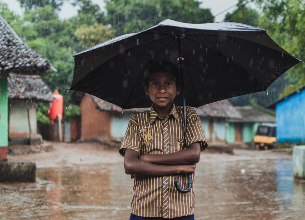 A young boy stands in the rain holding an umbrella in a village in Odisha, India.