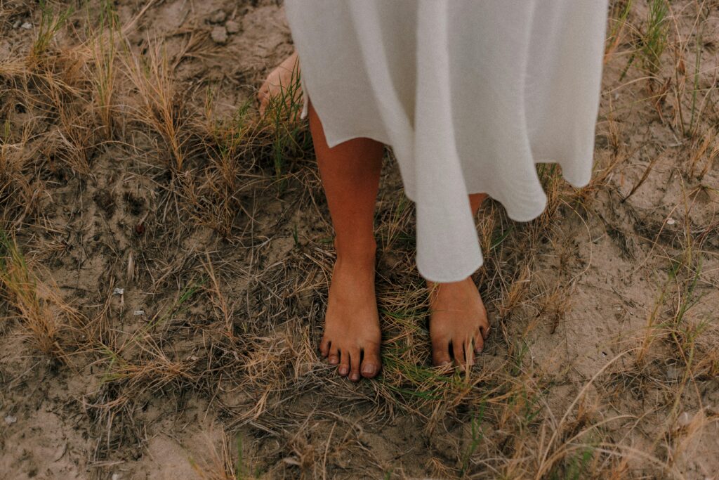 A woman dressed in a white garment stands barefoot on dry, sandy grass, creating a whimsical outdoor scene.
