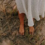 A woman dressed in a white garment stands barefoot on dry, sandy grass, creating a whimsical outdoor scene.
