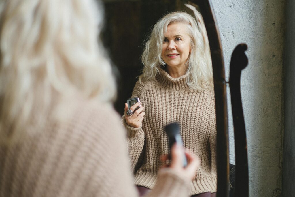 A senior woman in a cozy sweater smiles while applying makeup in front of a mirror.