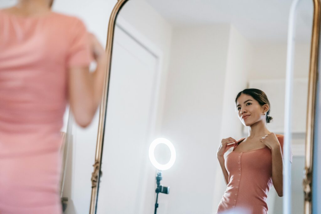 A young woman poses confidently in front of a mirror with a ring lamp.