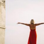 Woman in red dress with arms outstretched, enjoying freedom outdoors. Back view against a clear sky.