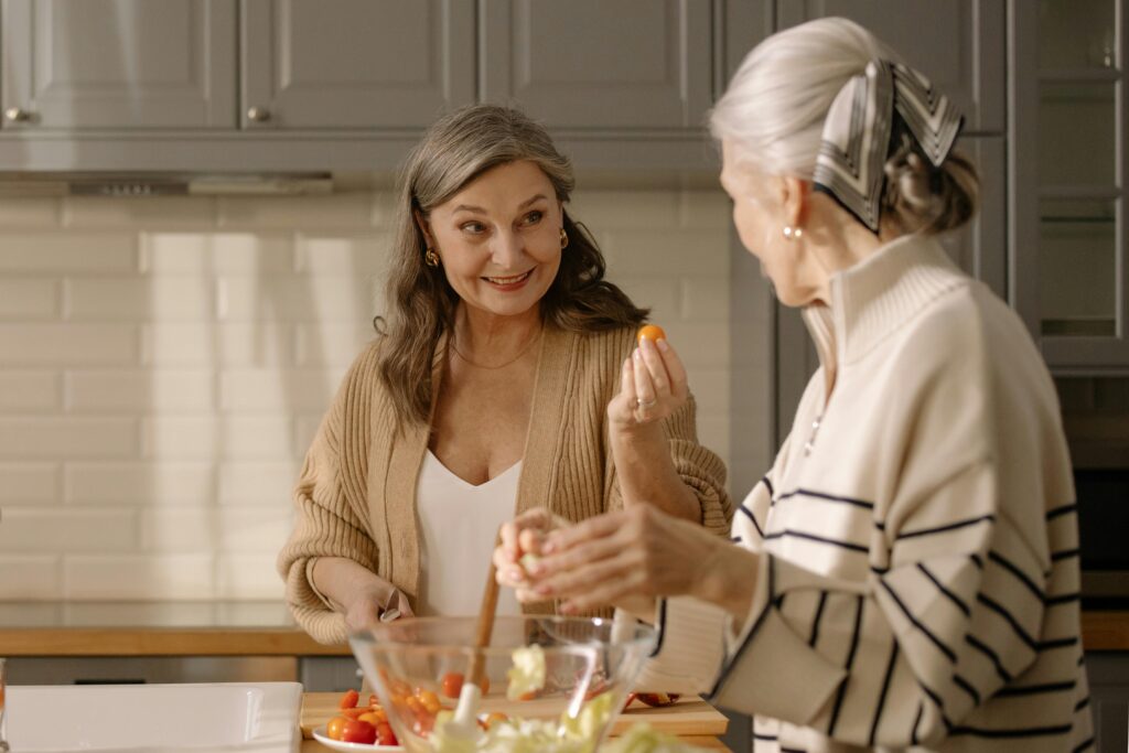 Two senior women smiling and preparing a fresh salad in a cozy kitchen setting, showcasing friendship and healthy living.