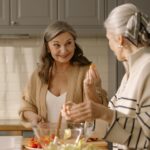 Two senior women smiling and preparing a fresh salad in a cozy kitchen setting, showcasing friendship and healthy living.