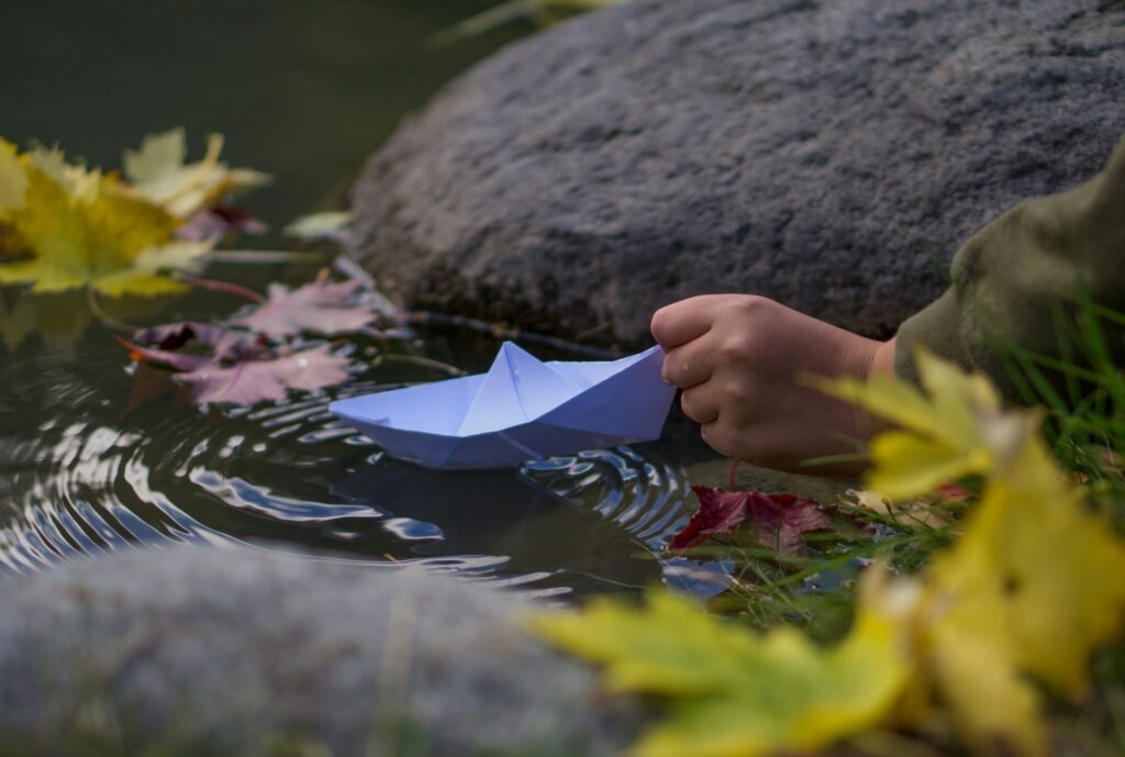Close-up of a child's hand launching a blue paper boat in a serene autumn stream surrounded by colorful leaves.
