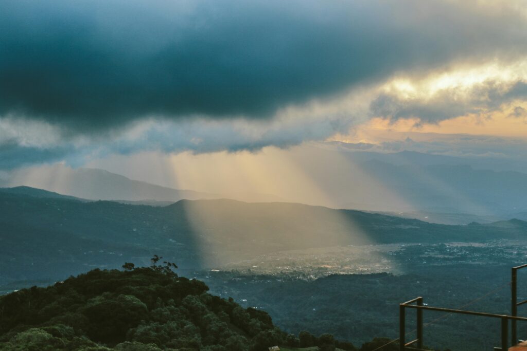 Sunbeams pierce through dramatic clouds over a lush mountain landscape, ideal for travel and nature themes.