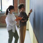 Two women collaborate at a blackboard in a classroom setting.