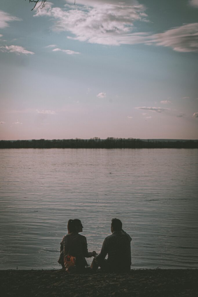 Silhouetted couple sitting by a calm lake under a serene sunset sky, enjoying the peaceful moment.