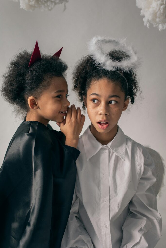 Two adorable children dressed as angel and devil whispering secrets during a Halloween studio shoot.