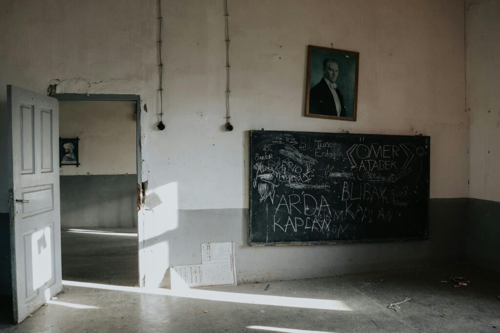 An empty, dimly lit classroom with an open door, blackboard, and portrait, evoking a historical atmosphere.