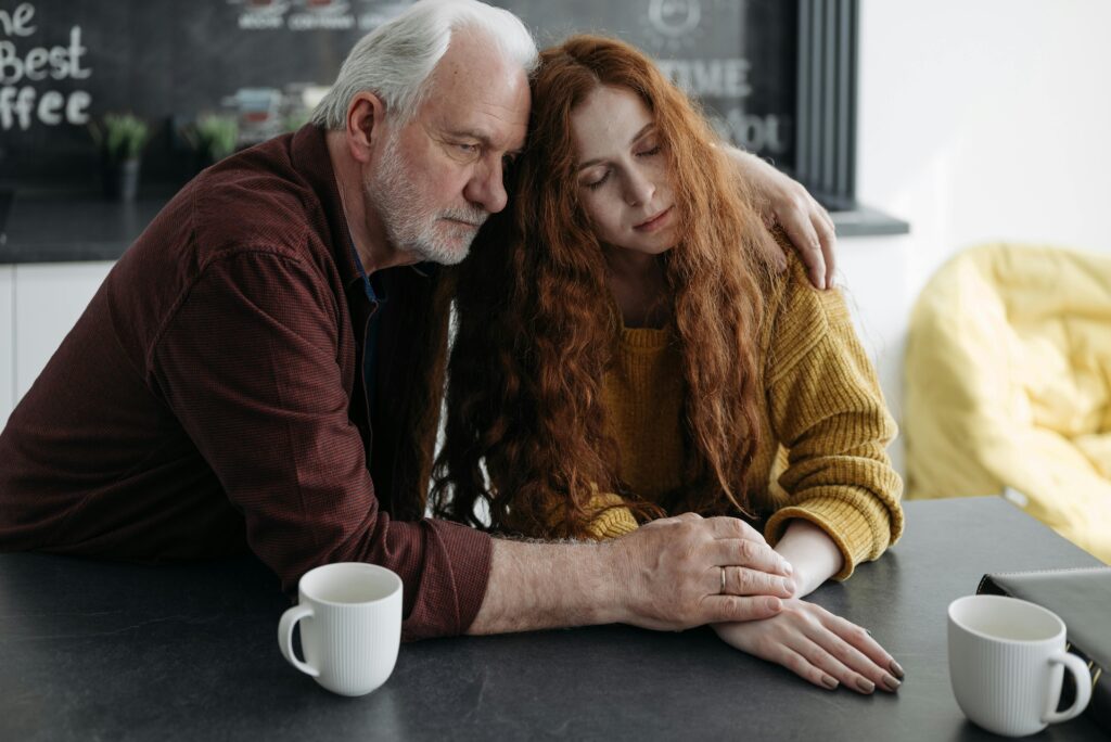 An elderly man consoles a young woman at a table in a warm, intimate indoor setting.