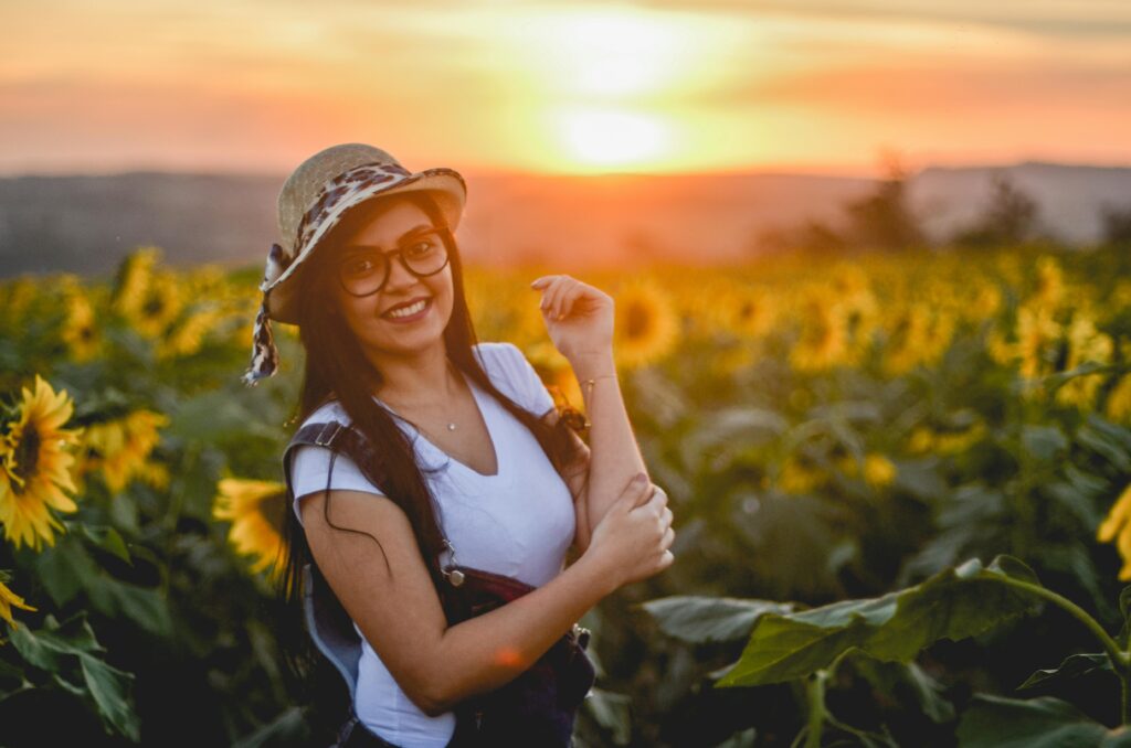 A woman wearing a straw hat and glasses smiling among sunflowers during sunset.