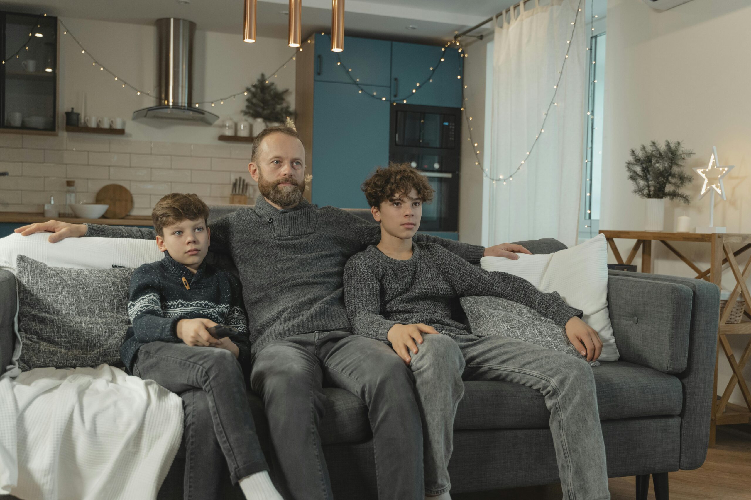 A father and his two sons sit comfortably on a sofa in a cozy, decorated living room.