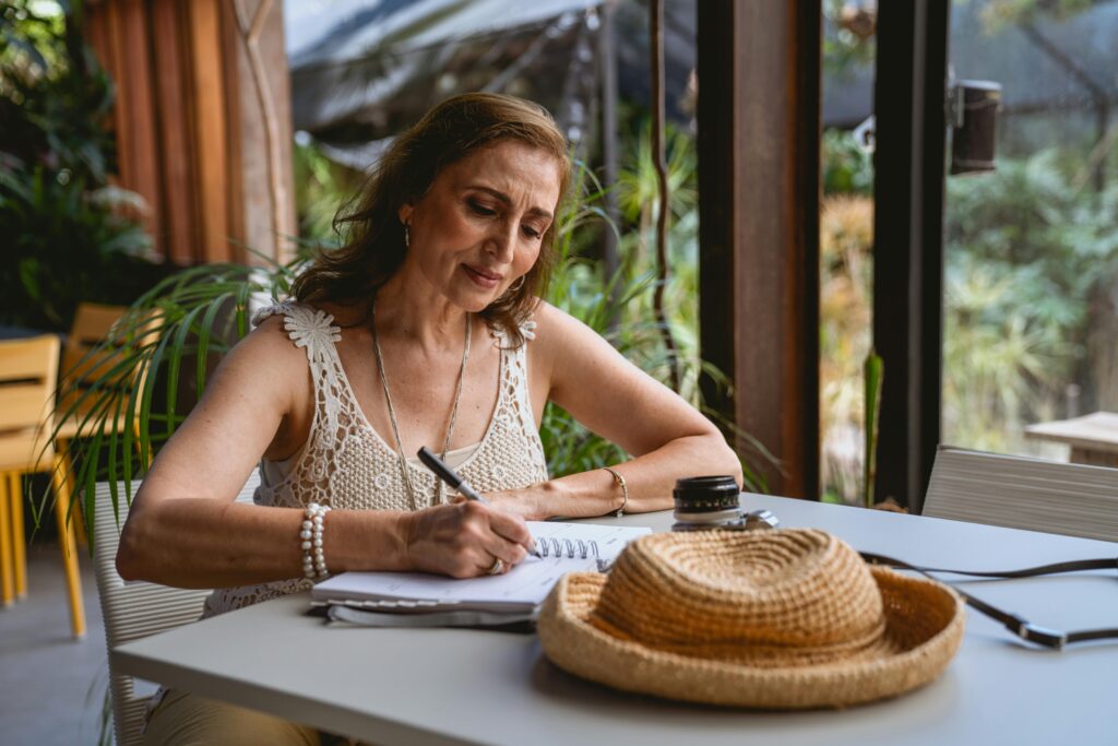 Elderly woman sitting at a cafe table, writing in a notebook with a straw hat nearby.