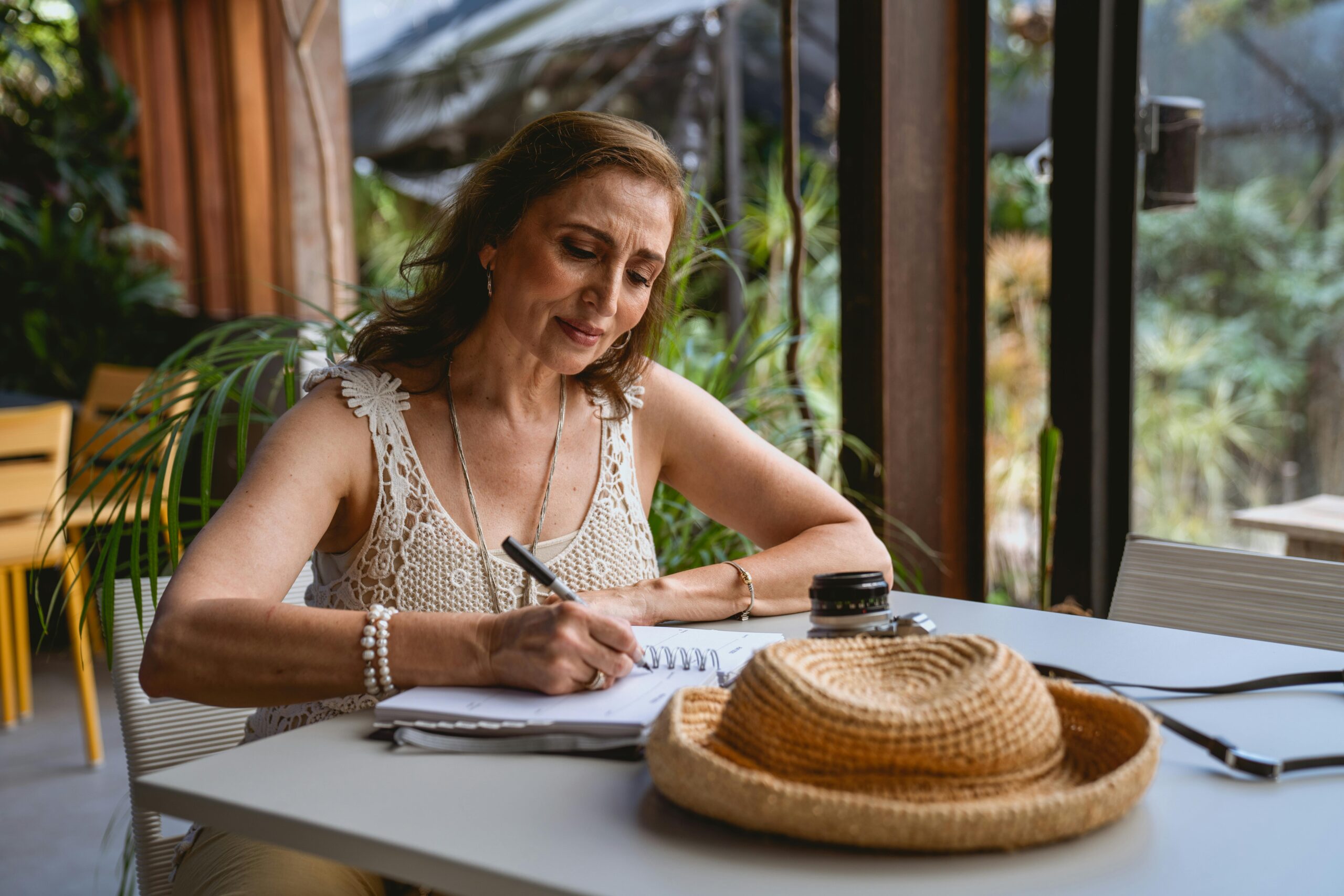 Elderly woman sitting at a cafe table, writing in a notebook with a straw hat nearby.