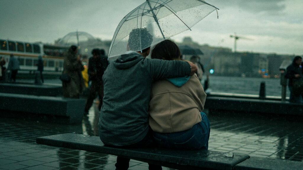 A couple sits close under an umbrella, embracing on a rainy city day.