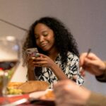 Happy young woman using smartphone while dining with friends indoors.
