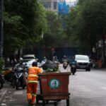 Street cleaner in orange uniform pushing a garbage bin on a city road, indicating urban sanitation.