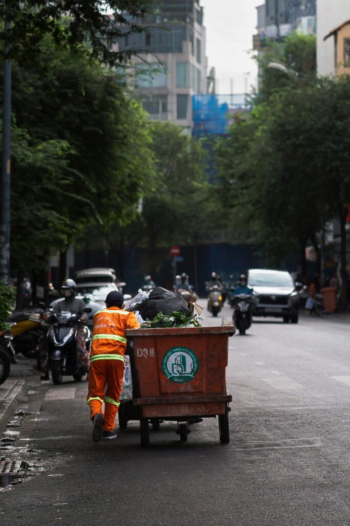 Street cleaner in orange uniform pushing a garbage bin on a city road, indicating urban sanitation.