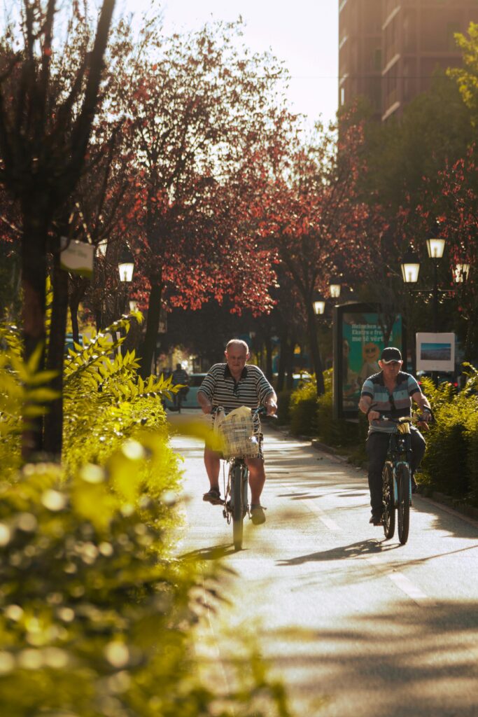 Two elderly men cycling on a sunny day through a tree-lined park pathway in Tirana, Albania.