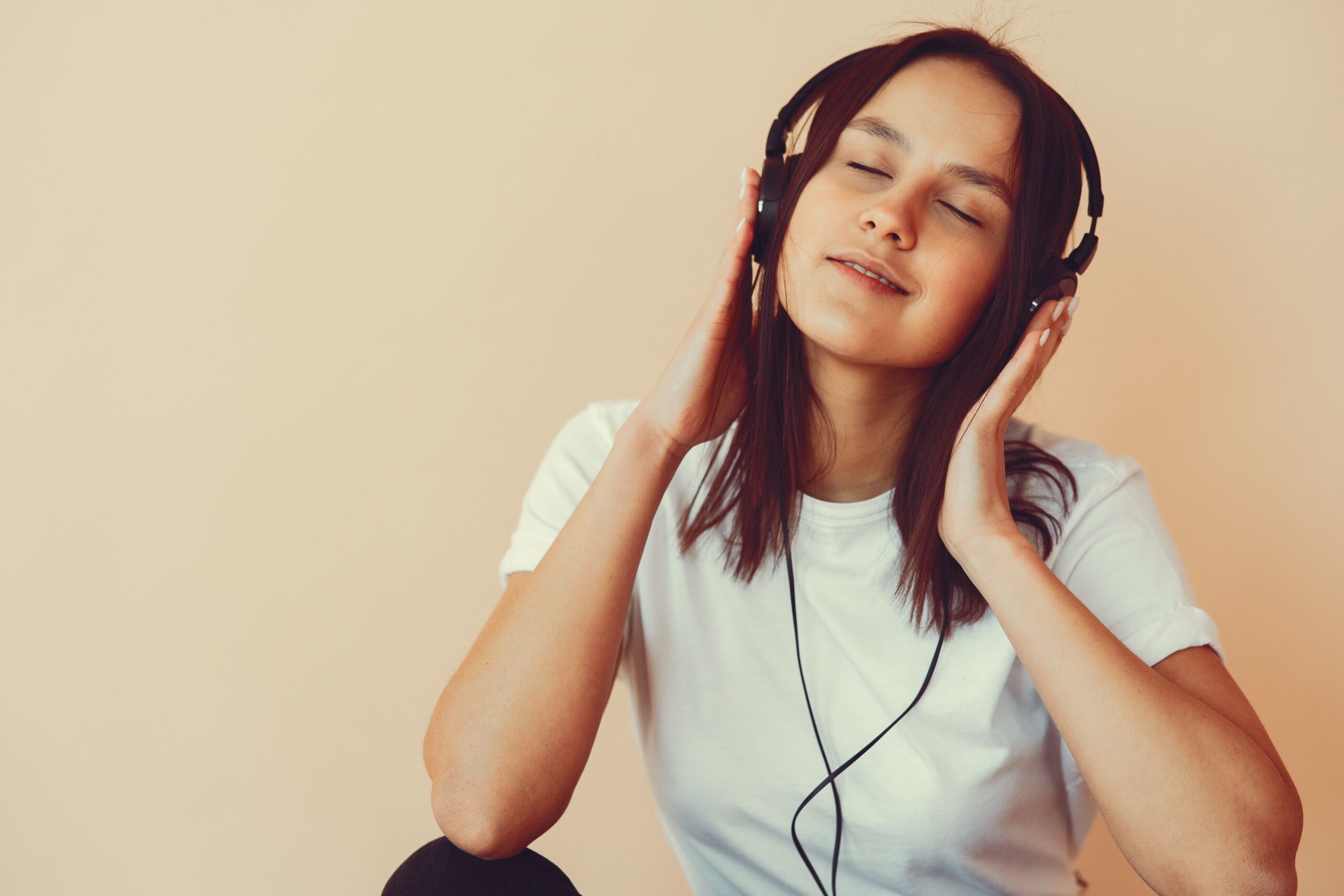 Positive young brunette in white shirt and headphones listening to good music and smiling happily while sitting and touching head against beige background