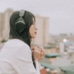 Side view of a woman in headphones and eyeglasses, enjoying a cityscape from a rooftop.
