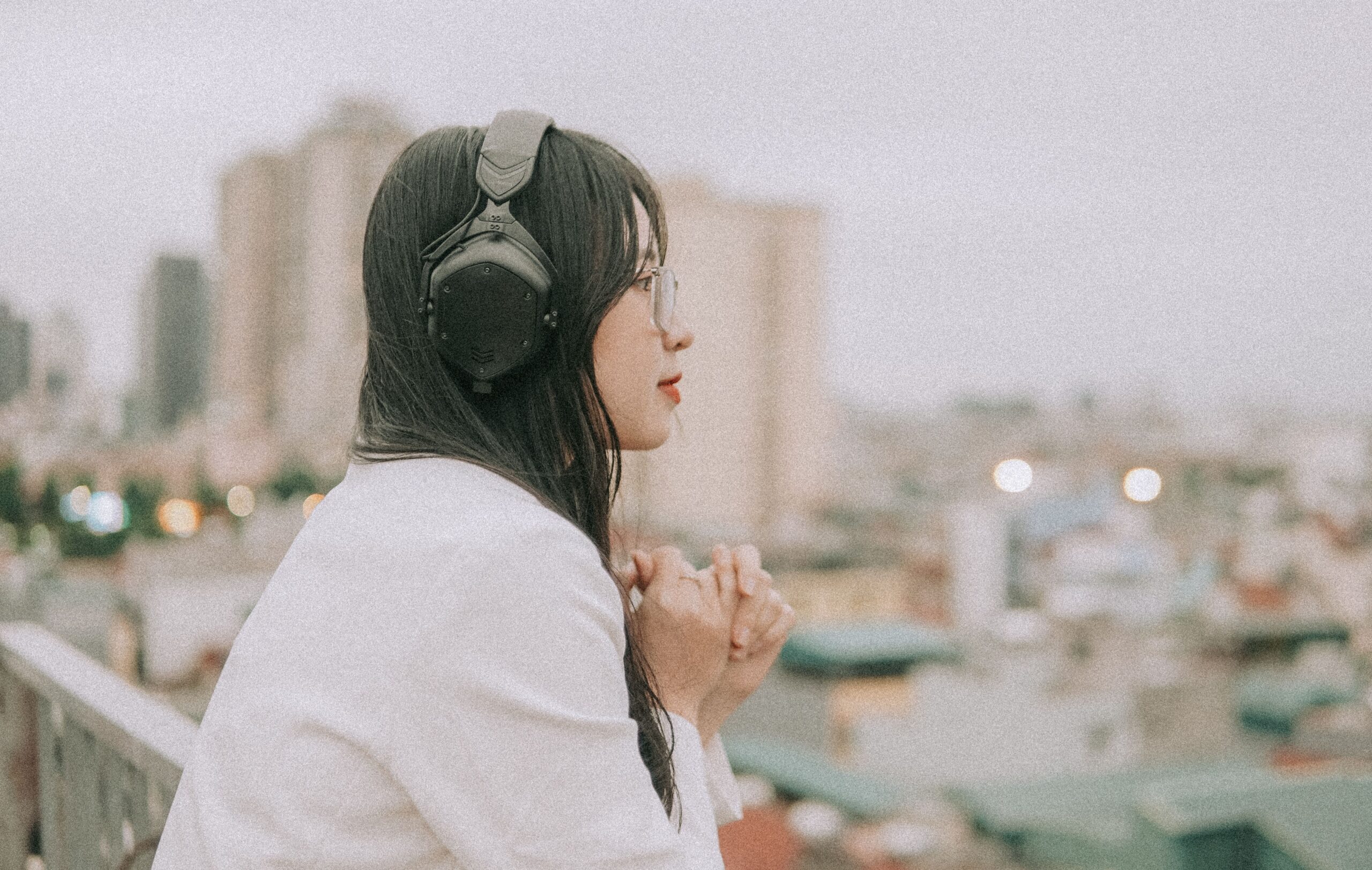Side view of a woman in headphones and eyeglasses, enjoying a cityscape from a rooftop.